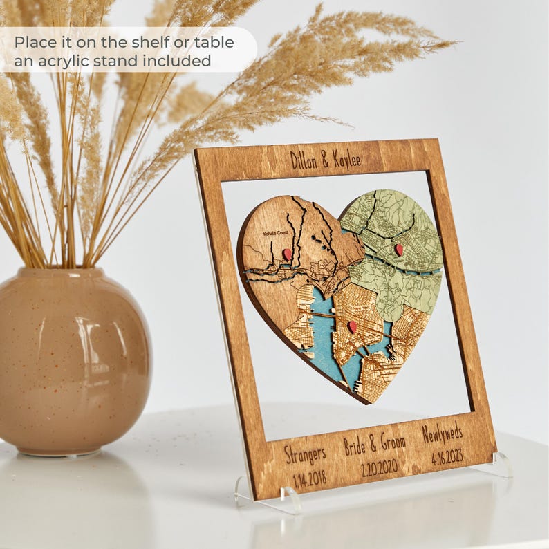 A warm wood framed heart puzzle map is displayed on an acrylic stand beside a ceramic vase, with text noting it can be placed on a shelf or table. This image shows the keepsake as a ready-made wedding gift for a couple that also works as home decor.