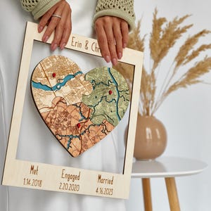 Angled close-up of the framed three-location heart puzzle map held in both hands beside a vase with pampas grass. This photo highlights the layered craftsmanship and romantic milestone layout of the personalized couple keepsake.