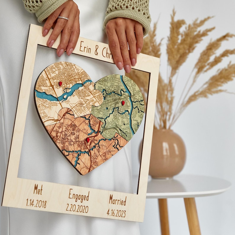 Angled close-up of the framed three-location heart puzzle map held in both hands beside a vase with pampas grass. This photo highlights the layered craftsmanship and romantic milestone layout of the personalized couple keepsake.