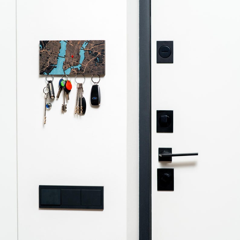 A compact custom magnetic key holder is mounted beside a modern front door with keys stored below the wooden map. The personalized location design adds function and meaning to a clean entryway.