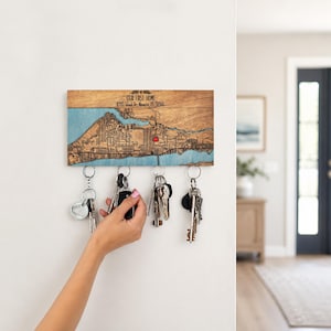 A personalized location map key holder is shown in a bright entryway while a hand reaches for keys. The wooden map organizer keeps daily keys easy to grab near the front door.