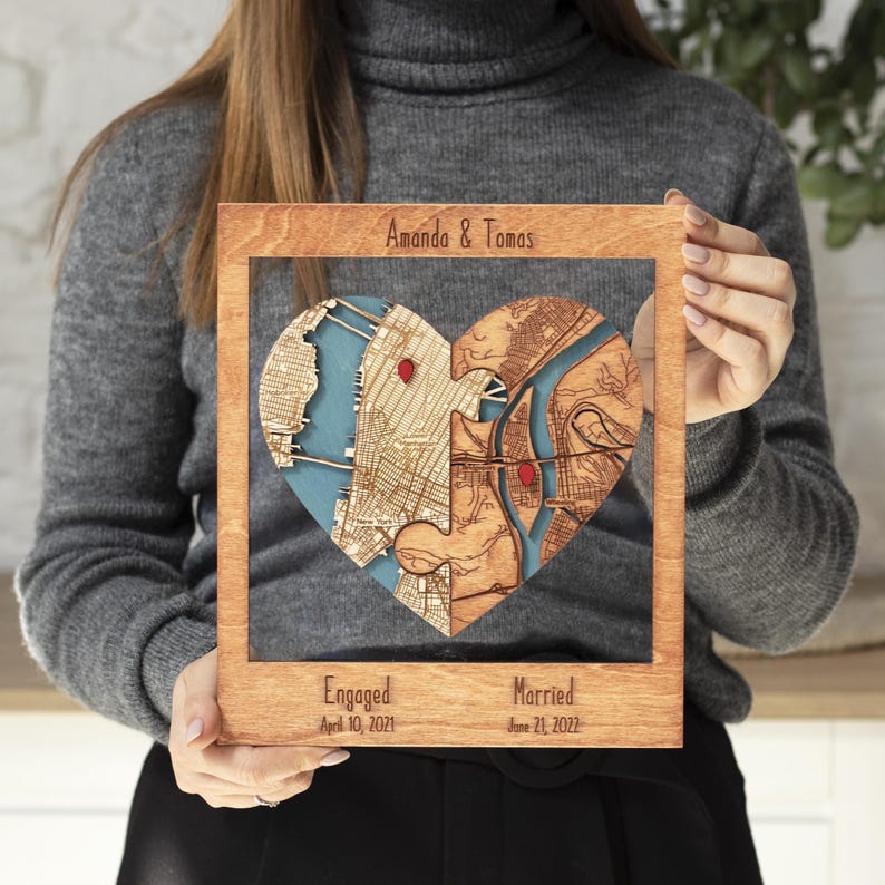A woman holds a framed wooden heart map personalized for a couple, with two joined locations and milestone dates below. The image presents the piece as a sentimental engagement or wedding keepsake designed for wall or shelf display.