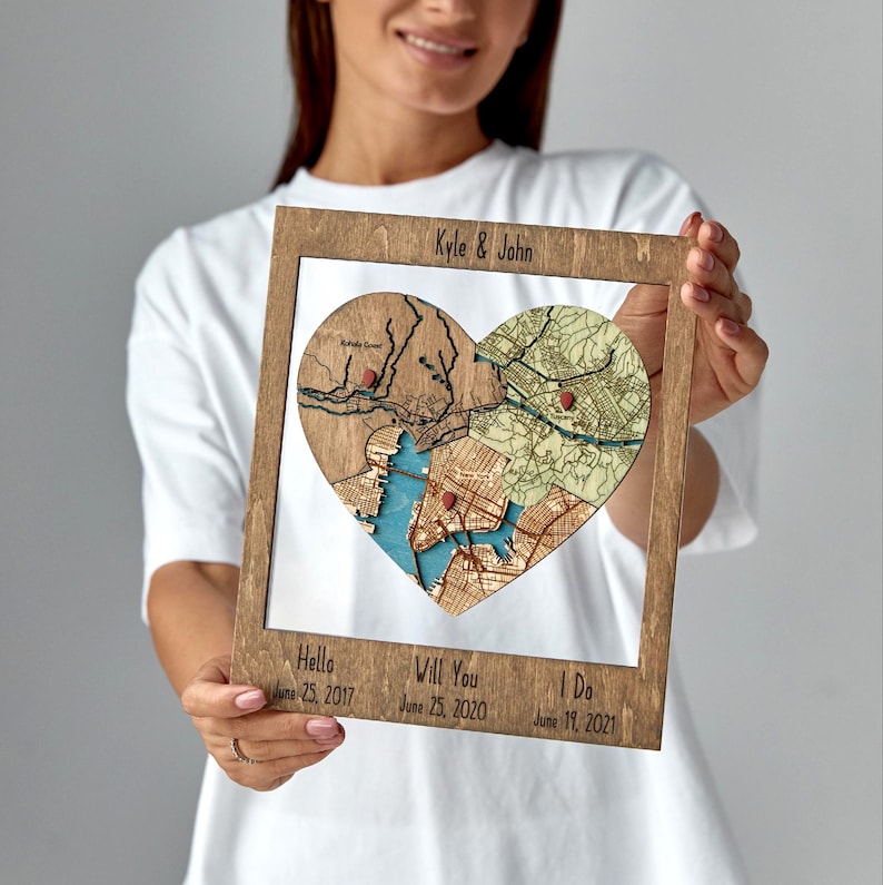 Framed wooden wall art featuring a heart-shaped puzzle of layered location maps with red pins, personalized with couple names on top and dates on the bottom.
