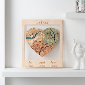 May include: A wooden heart-shaped map with three sections, framed in light wood. The map shows different locations with red pin markers. The top of the frame reads "Erin & Chris", and the bottom has dates for "Met", "Engaged", and "Married".