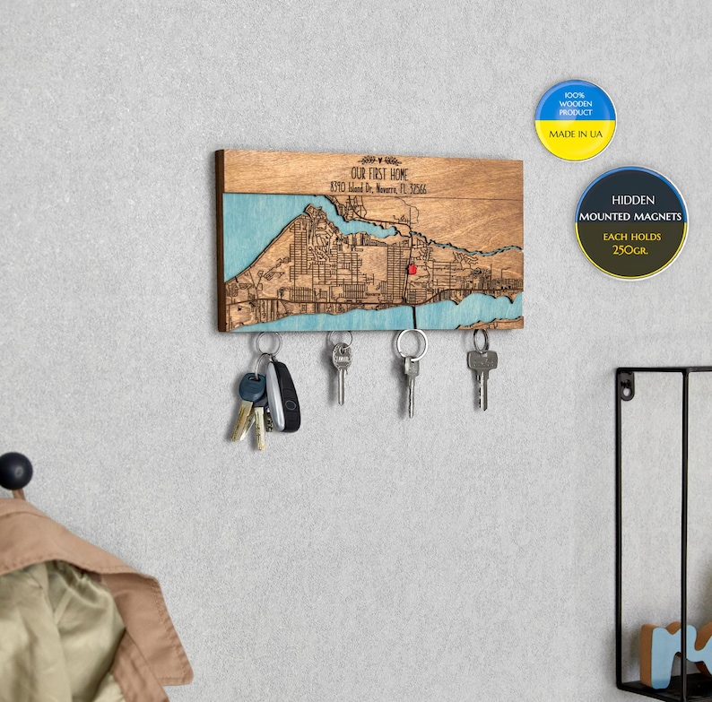 May include: Wooden key holder featuring a map of a city with the inscription "OUR FIRST HOME 8390 Island Dr. New, FL 32566". The key holder has five hooks for keys and hidden magnets to keep them secure. The key holder is crafted from wood and has a blue and brown colour scheme.