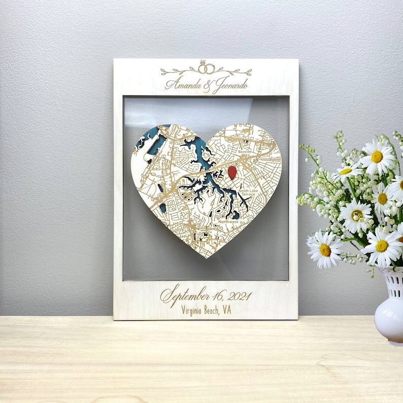 May include: A white wooden frame with a heart-shaped map of Virginia Beach, Virginia. The map is laser-cut and features a teal blue water color. The frame includes the names "Amanda & Leonardo" and the date "September 16, 2021".