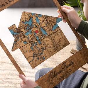 A person holds the house-shaped wooden map inside the frame, showing the detailed map design up close. This photo emphasizes the custom address area and the handcrafted quality of the personalized first home gift.