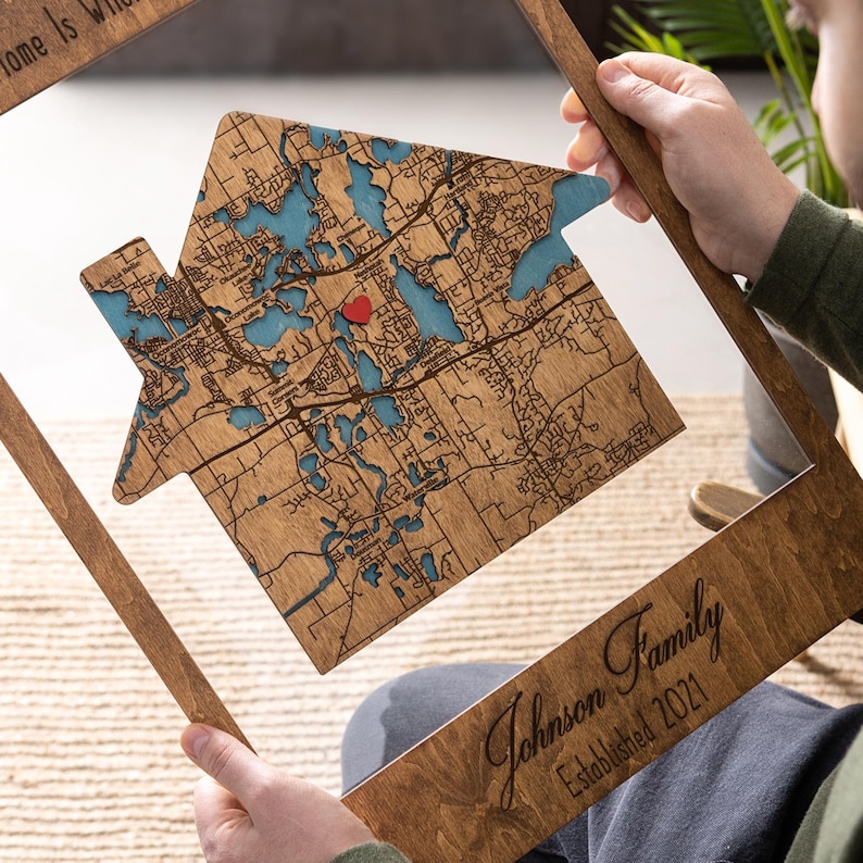 A person holds the house-shaped wooden map inside the frame, showing the detailed map design up close. This photo emphasizes the custom address area and the handcrafted quality of the personalized first home gift.