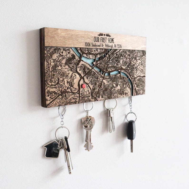 May include: A wooden key holder featuring a laser-engraved map of Pittsburgh, Pennsylvania. The holder is inscribed with the text "OUR FIRST HOME" and an address. Five keys are hanging from the holder.