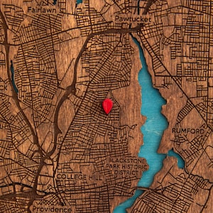 May include: A wooden map of Providence, Rhode Island, featuring laser-cut street grids in dark brown wood with a bright turquoise water feature. A small, solid red teardrop-shaped location pin is placed on the map surface to mark a specific point.