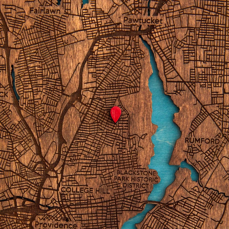 May include: A wooden map of Providence, Rhode Island, featuring laser-cut street grids in dark brown wood with a bright turquoise water feature. A small, solid red teardrop-shaped location pin is placed on the map surface to mark a specific point.