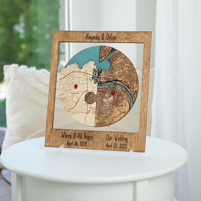 Personalized wooden circle map displayed on a white side table with names and milestone captions marking where it all began and the wedding date. This styling presents the piece as a meaningful shelf keepsake for a couple’s engagement or wedding story.