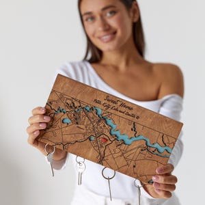 May include: A wooden map with a city layout, featuring a river in blue resin. The map has the words "Sweet Home" and a name inscription. Keys hang from the bottom. The map is held by a person.