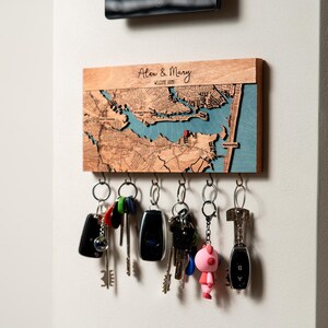 Close-up of a personalized wooden key rack with engraved map detail, couple names, and multiple hanging keys. This image emphasizes the decorative map surface and the practical hook layout for daily entryway use.
