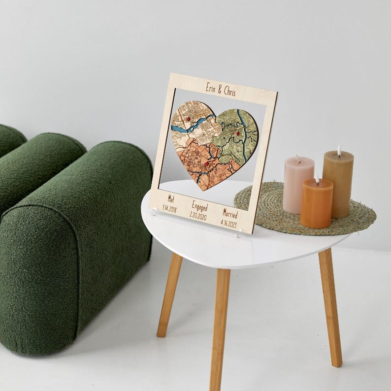 A light wood framed heart puzzle map stands on a small round table beside candles and a green chair. This image presents the personalized piece as elegant shelf or tabletop decor for celebrating a couple’s love story.