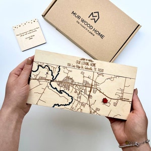 May include: A wooden map of Gatesville, TX, with a red marker, held in hands. The map is engraved with streets and a river. Text on the map reads "OUR LOVING HOME" and the address. A small wooden card and a box with the text "MUR WOOD HOME" are also visible.