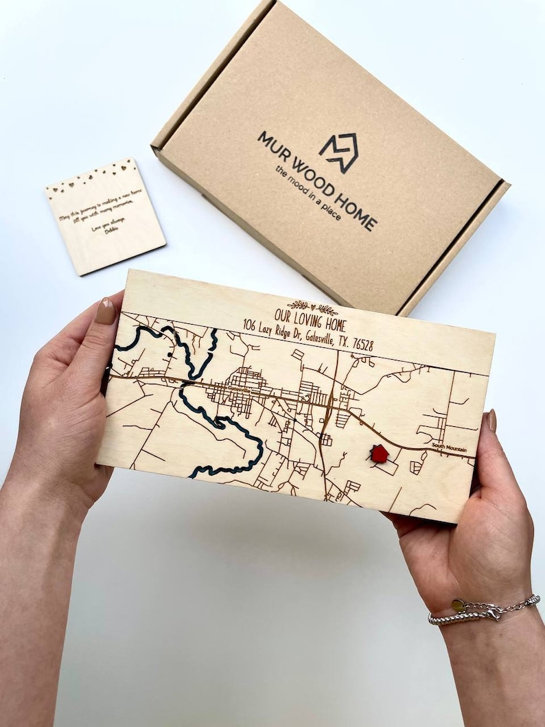May include: A wooden map of Gatesville, TX, with a red marker, held in hands. The map is engraved with streets and a river. Text on the map reads "OUR LOVING HOME" and the address. A small wooden card and a box with the text "MUR WOOD HOME" are also visible.