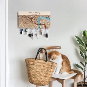 A personalized map key holder is mounted above a woven bag beside a cat in a cozy entryway corner. The wooden magnetic organizer adds practical key storage and warm home decor.