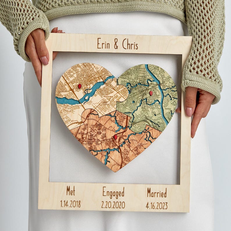 A person holds a light wood framed heart puzzle map personalized with names and milestone dates for where the couple met, got engaged, and married. This image presents the piece as a sentimental personalized wedding gift for a couple with three meaningful locations in one keepsake.