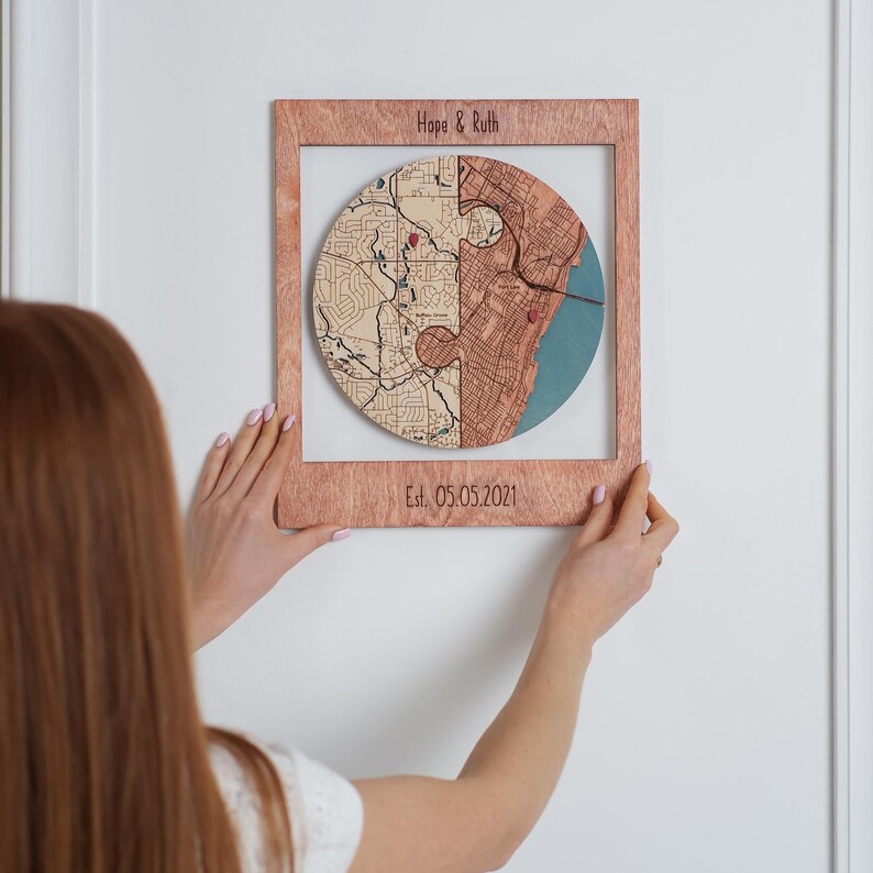 A woman hangs a personalized wooden circle map on the wall, showing the artwork as ready-to-display decor for a couple’s shared story. This image highlights the custom map keepsake as romantic wall art for an engagement, anniversary, or wedding gift.