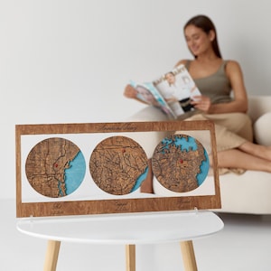 May include: A rectangular wooden frame containing three circular laser-cut maps with blue water details and red location markers. The frame features the text "Amanda and Tierney" at the top and "Met", "Engaged 10.2.2022", and "Married 10.14.2023" below.