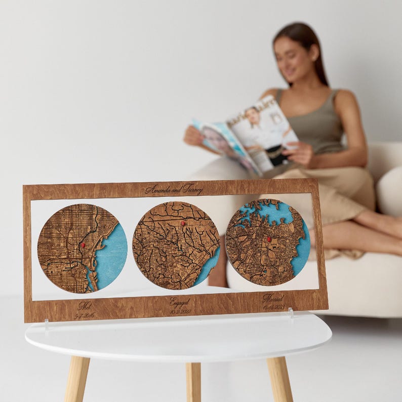 May include: A rectangular wooden frame containing three circular laser-cut maps with blue water details and red location markers. The frame features the text "Amanda and Tierney" at the top and "Met", "Engaged 10.2.2022", and "Married 10.14.2023" below.