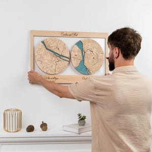 May include: A rectangular wooden wall art piece featuring two circular map inlays with blue water details and red location markers. The frame includes the text Caroline and Matt, Where it all began, and a date. The wood has a light natural finish.