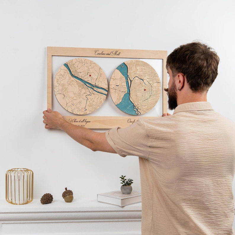 May include: A rectangular wooden wall art piece featuring two circular map inlays with blue water details and red location markers. The frame includes the text Caroline and Matt, Where it all began, and a date. The wood has a light natural finish.