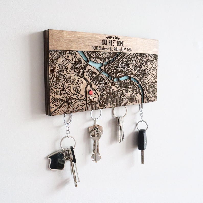 A personalized location map key holder is mounted on a white wall with several key sets held below by hidden magnets. This custom wooden entryway organizer makes a meaningful housewarming gift or realtor closing gift.