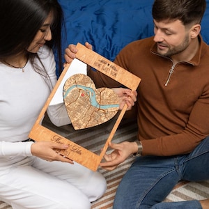 A couple sits together holding a framed heart-shaped map, showing a warm and personal version of this custom first home gift. The image highlights the romantic, giftable feel of the personalized wood wall art.