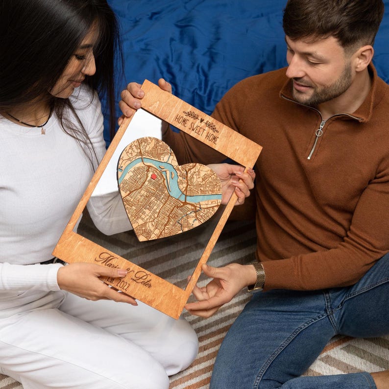 A couple sits together holding a framed heart-shaped map, showing a warm and personal version of this custom first home gift. The image highlights the romantic, giftable feel of the personalized wood wall art.