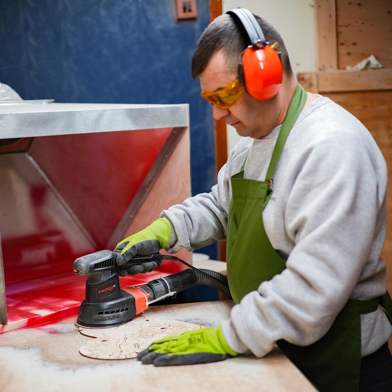 A maker sands a wooden heart puzzle map piece in the workshop while wearing protective gear. This image emphasizes the handmade process and craftsmanship behind the personalized couple gift.