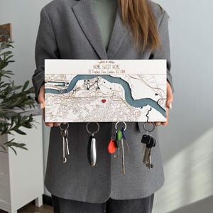 A person holds a white wooden magnetic key organizer personalized with “Home Sweet Home” and a custom location map. The key holder combines entryway organization with a meaningful home address design.