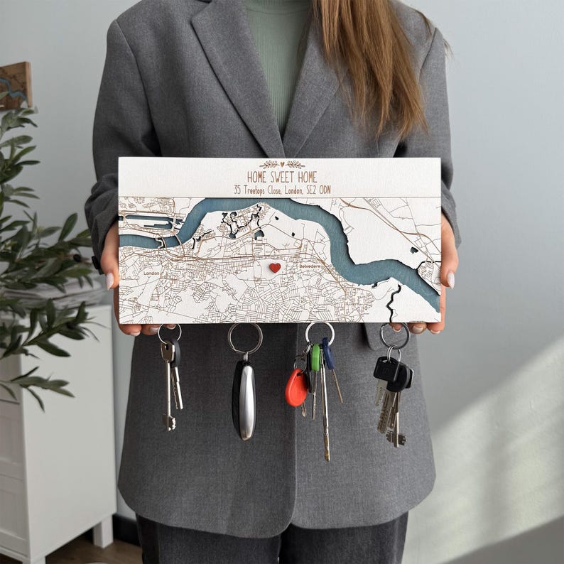 A person holds a white wooden magnetic key organizer personalized with “Home Sweet Home” and a custom location map. The key holder combines entryway organization with a meaningful home address design.