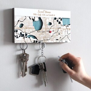 May include: A wooden key holder with a map design, featuring the text "Sweet Home" and names. The map has a heart marking a location. Several keys hang from the key holder. A hand is reaching for a key.