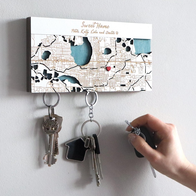 May include: A wooden key holder with a map design, featuring the text "Sweet Home" and names. The map has a heart marking a location. Several keys hang from the key holder. A hand is reaching for a key.