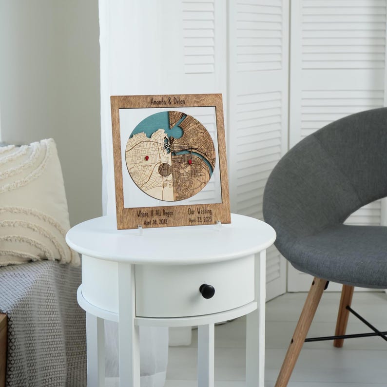 Wooden circle map keepsake styled on a white side table in a bright room with soft furnishings and a chair nearby. The image helps buyers picture the personalized engagement and wedding gift as elegant shelf decor in a modern home.