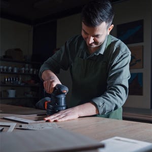 May include: A person wearing a green apron and a long-sleeved shirt uses a black and orange electric orbital sander to smooth the surface of a flat wooden frame on a workbench in a workshop setting with dim lighting.