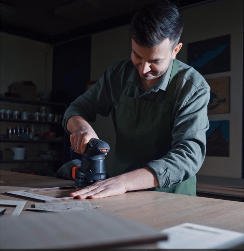 May include: A person wearing a green apron and a long-sleeved shirt uses a black and orange electric orbital sander to smooth the surface of a flat wooden frame on a workbench in a workshop setting with dim lighting.