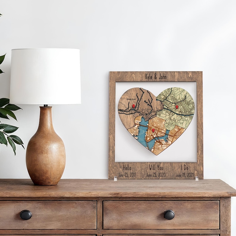A framed heart puzzle map stands on a dresser beside a lamp and plant, personalized with names and milestone dates from hello to proposal to marriage. This image presents the keepsake as refined romantic decor for a couple’s home.