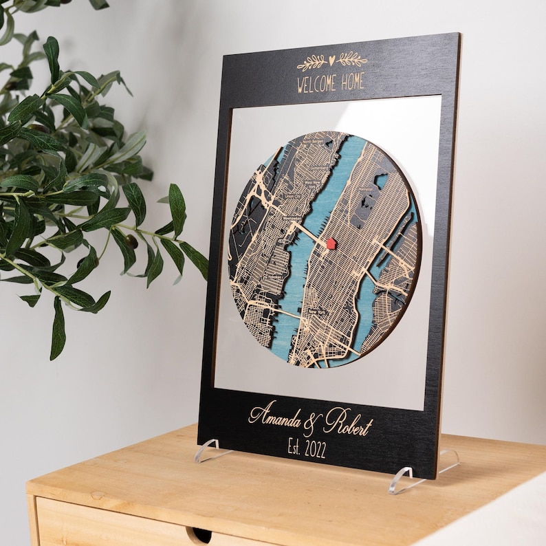 A black-framed round map stands on a tabletop stand, showing a personalized housewarming gift for a new home. This photo highlights the clean display style, layered wood details, and custom engraved names and date.