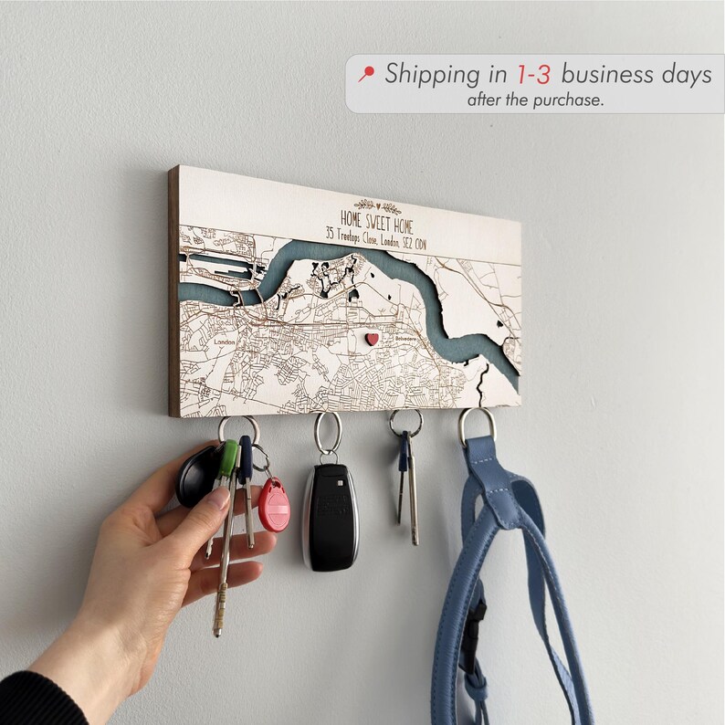 A white personalized location map key holder is mounted on a wall with keys and a small bag held below. The image highlights fast processing and a clean custom design for a new home gift.