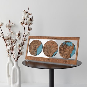 May include: A rectangular wooden frame containing three circular laser-cut maps with blue water details. The frame features the text Amanda and Jeremy at the top, with Met, Engaged, and Married printed below each map along with specific dates.