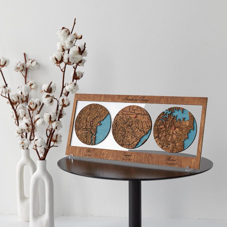 May include: A rectangular wooden frame containing three circular laser-cut maps with blue water details. The frame features the text Amanda and Jeremy at the top, with Met, Engaged, and Married printed below each map along with specific dates.