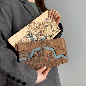 May include: Two wooden map art pieces, one light and one dark, are held up. The light wood piece says "OUR FIRST HOME" and the address. The dark wood piece says "HOME SWEET HOME" and the address. Both have a blue river and a red heart.