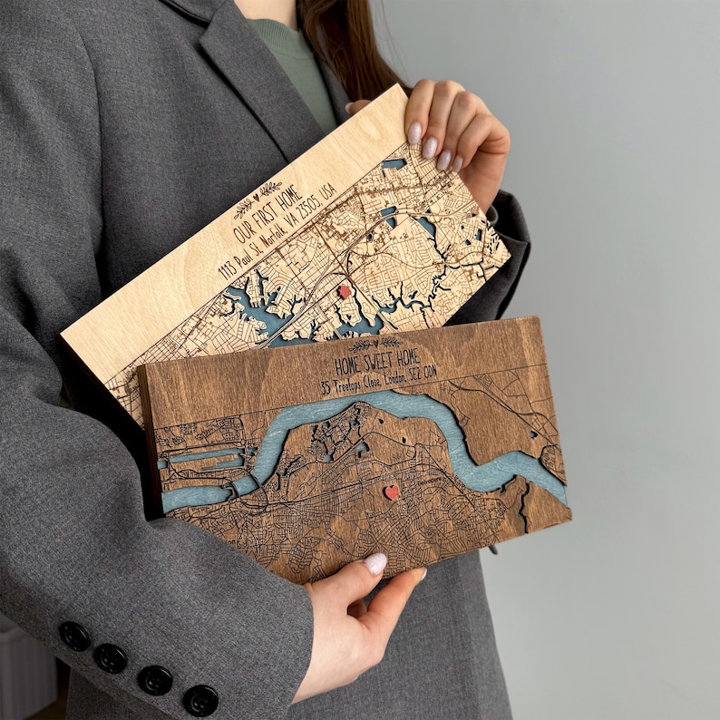 May include: Two wooden map art pieces, one light and one dark, are held up. The light wood piece says "OUR FIRST HOME" and the address. The dark wood piece says "HOME SWEET HOME" and the address. Both have a blue river and a red heart.