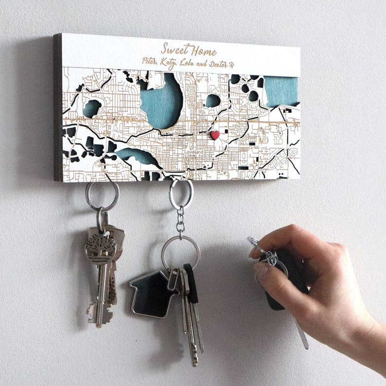 May include: A white wooden key holder with a map of a city and the text "Sweet Home Peter, Katy, Lola and Dexter" on it. There are three keychains hanging from the holder.