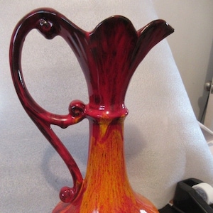 May include: A red and orange ceramic pitcher with a flared spout and a decorative handle. The pitcher has a glossy finish and a unique glaze pattern.