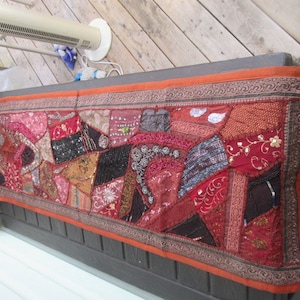 May include: A long, rectangular tapestry with a patchwork design. The tapestry is made of various fabrics in shades of red, brown, black, and gold. The fabric is decorated with sequins, beads, and embroidery. The tapestry has an orange border with a brown and gold design.