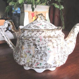 May include: A vintage white porcelain teapot with a gold floral pattern and accents. The teapot has a curved spout, a decorative handle, and a lid with a gold finial. Pink and green floral accents are scattered throughout the design. The teapot sits on a white base.